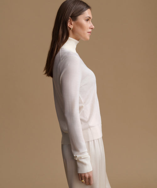 A woman in side profile wearing a cream-colored mock neck knit sweater and matching skirt against a solid beige background.