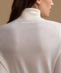 Close-up back view of a woman wearing a cream-colored ribbed turtleneck sweater against a warm beige background.
