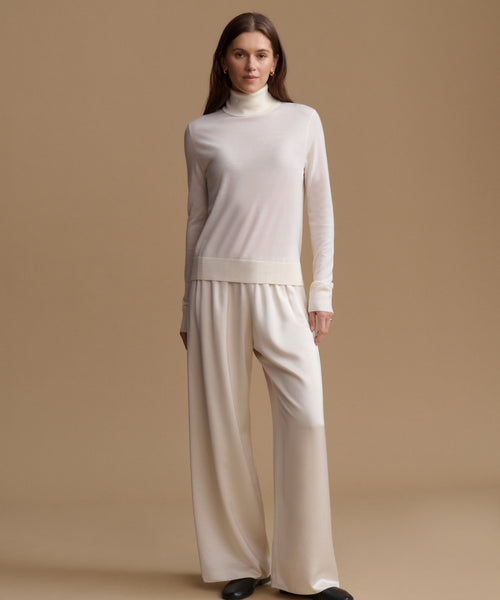 A woman modeling a cream-colored knit turtleneck sweater and matching wide-leg trousers against a solid beige studio background.