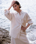 Wearing the Linen Colby Shirt, a person with short dark hair stands by the water, striking a thoughtful pose—one hand on their head and one on their hip—for an effortlessly stylish summer look.