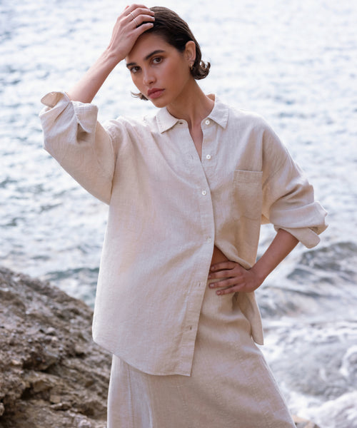Wearing the Linen Colby Shirt, a person with short dark hair stands by the water, striking a thoughtful pose—one hand on their head and one on their hip—for an effortlessly stylish summer look.