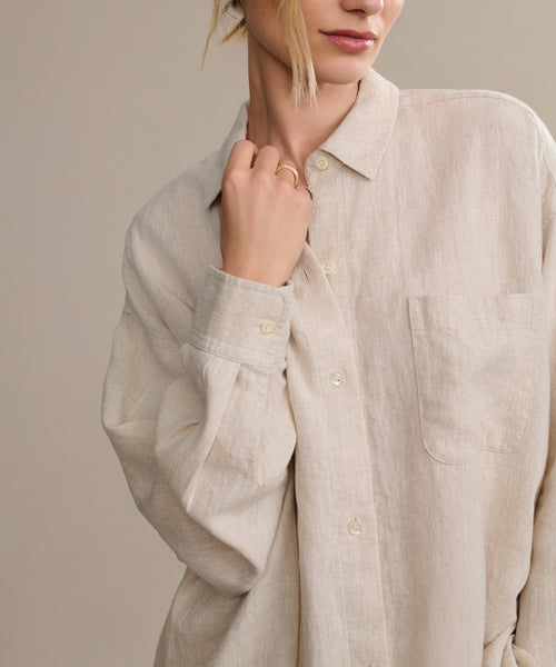 A person wears the loose, beige Linen Colby Shirt with a chest pocket, holding the collar with one hand. Their face is partly visible and hair tied back—ideal for relaxed summer style.