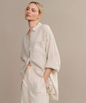 A woman with blonde hair in a low bun models the oversized Linen Colby Shirt in light beige with rolled sleeves, paired with cream pants. She stands against a neutral backdrop, one hand in her pocket—a perfect summer style.