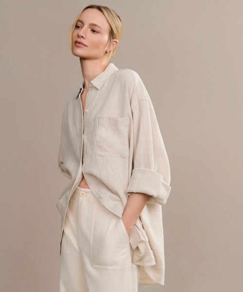 A woman with blonde hair in a low bun models the oversized Linen Colby Shirt in light beige with rolled sleeves, paired with cream pants. She stands against a neutral backdrop, one hand in her pocket—a perfect summer style.