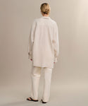 A person stands with their back to the camera, wearing the oversized Linen Colby Shirt in light beige, paired with loose matching pants and dark sandals, hair neatly tied in a bun against a plain backdrop.