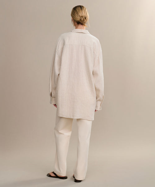 A person stands with their back to the camera, wearing the oversized Linen Colby Shirt in light beige, paired with loose matching pants and dark sandals, hair neatly tied in a bun against a plain backdrop.