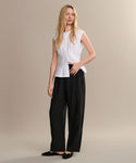A woman with long blonde hair and black flats stands against a beige background, modeling the Linen Logan Pant in black with a sleeveless white blouse, embodying effortless everyday style.