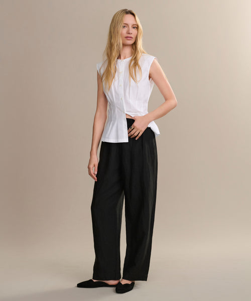 A woman with long blonde hair and black flats stands against a beige background, modeling the Linen Logan Pant in black with a sleeveless white blouse, embodying effortless everyday style.