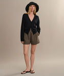A woman in a black wide-brim hat, black V-neck cardigan, high-waisted brown Linen Logan Short, and black sandals stands against a plain light beige background.