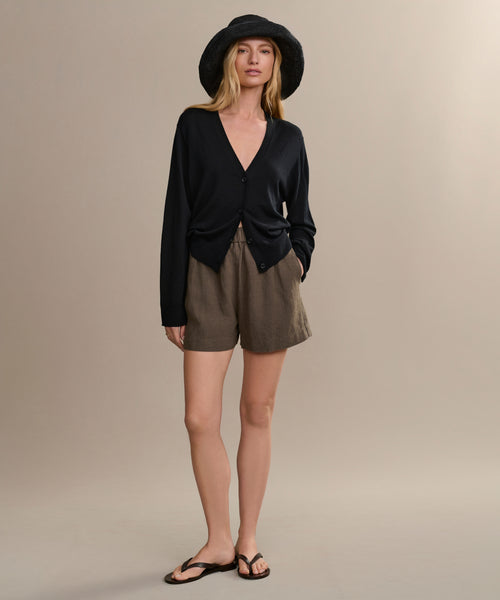 A woman in a black wide-brim hat, black V-neck cardigan, high-waisted brown Linen Logan Short, and black sandals stands against a plain light beige background.