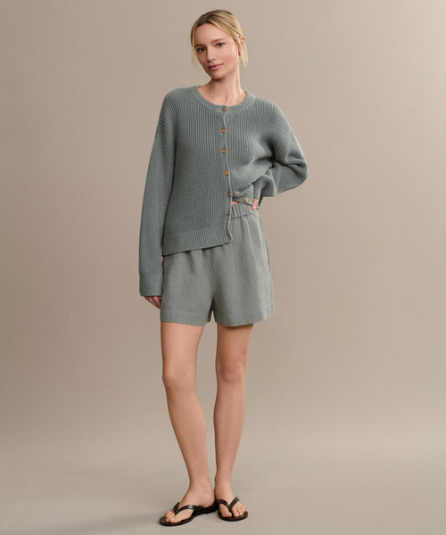 A woman with blonde hair pulled back stands against a plain beige background, wearing the Linen Logan Short, a light gray ribbed cardigan, and black sandals—capturing an easy, breezy warm-weather style.