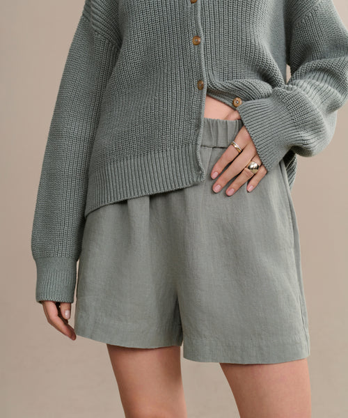 A person models warm-weather style in a loose, textured sage green cardigan with brown buttons and Linen Logan Shorts. Only the torso and upper legs are visible as their ring-adorned hand rests on their hip.