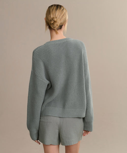 A person with blonde hair in a low bun models the Linen Logan Short in gray-green, styled for warm weather, standing with their back to the camera against a plain beige background.