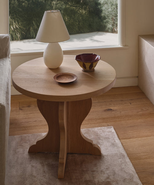 The Lombard Side Table in solid white oak holds a white lamp, wooden dish, and red ceramic bowl. Placed on a beige rug by the sofa, it’s set against a window with lush greenery in the background.
