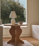 The Lombard Side Table, made of solid white oak, displays a white lamp and decorative bowl near a large window with sunlight streaming in and greenery outside.