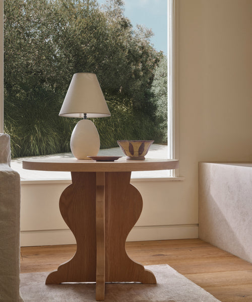 The Lombard Side Table, made of solid white oak, displays a white lamp and decorative bowl near a large window with sunlight streaming in and greenery outside.