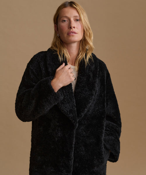A woman wearing a black shearling coat over a light beige top, standing against a solid beige background.