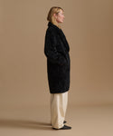 A woman stands in profile wearing a long black shearling coat over cream wide-leg trousers and black pointed shoes, photographed against a plain warm beige studio background.