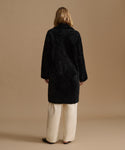 A model viewed from the back wearing a black shearling coat over cream wide-leg trousers, set against a solid warm beige studio background.