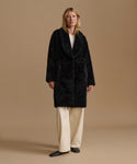 A woman stands against a plain beige studio background wearing a black shaggy shearling coat over a white top and cream wide-leg trousers.