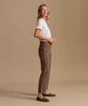 A woman stands in profile wearing brown straight-leg trousers, a white short-sleeve top, and dark loafers against a plain beige studio background.