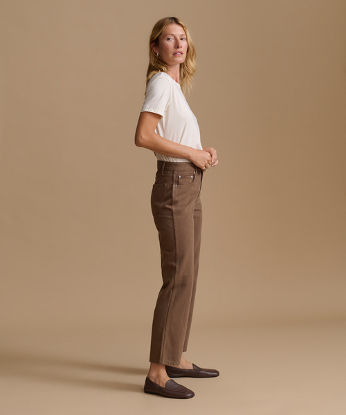 A woman stands in profile wearing brown straight-leg trousers, a white short-sleeve top, and dark loafers against a plain beige studio background.
