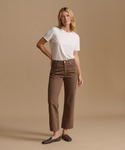 A woman stands against a plain beige studio background wearing a white short-sleeve T-shirt, brown cropped pants, and dark brown slip-on shoes.