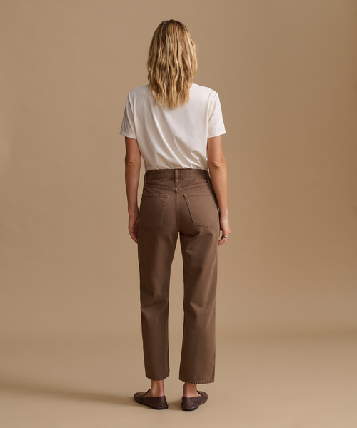A woman viewed from the back wearing brown straight-leg cropped pants, a white t-shirt, and dark brown loafers, set against a solid beige studio background.