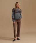 A woman stands against a plain beige studio background wearing a gray knit cardigan with a brown and cream Fair Isle yoke pattern, brown cropped pants, and dark loafers.