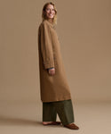A woman modeling a long tan coat over olive green wide-leg pants and brown suede shoes, standing against a solid tan studio background.