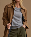 A woman wearing a brown trench coat over a heather gray crewneck sweater and olive green trousers, shown from the neck down against a solid warm beige studio background.