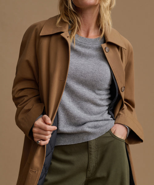 A woman wearing a brown trench coat over a heather gray crewneck sweater and olive green trousers, shown from the neck down against a solid warm beige studio background.