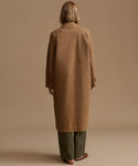 A woman viewed from behind wearing a long camel brown coat over olive green trousers, standing against a solid beige studio background.