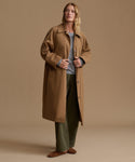 A woman stands in a studio wearing a long tan trench coat over a light gray top and olive green pants, against a warm beige background.