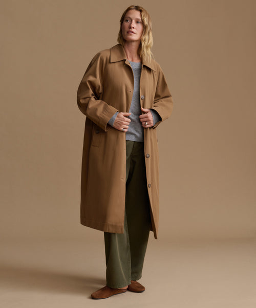 A woman stands in a studio wearing a long tan trench coat over a light gray top and olive green pants, against a warm beige background.