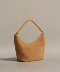 The Medium Crescent Bag in camel-colored suede features a curved design and thick twisted handle, crafted in Italy. Photographed on a plain beige background.