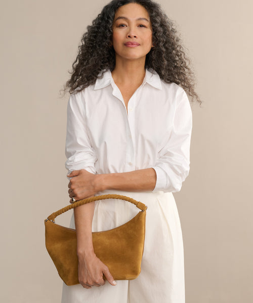A woman in a white button-up shirt and pants holds the Medium Crescent Bag, crafted in Italy. The bag’s tan suede finish stands out against a soft beige background.