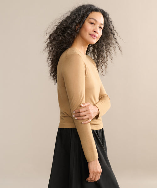 A woman with long, curly hair models the Mercer Crewneck in a stylish look, paired with a black skirt. She stands sideways against a plain beige background, smiling gently with one arm crossed over her body.
