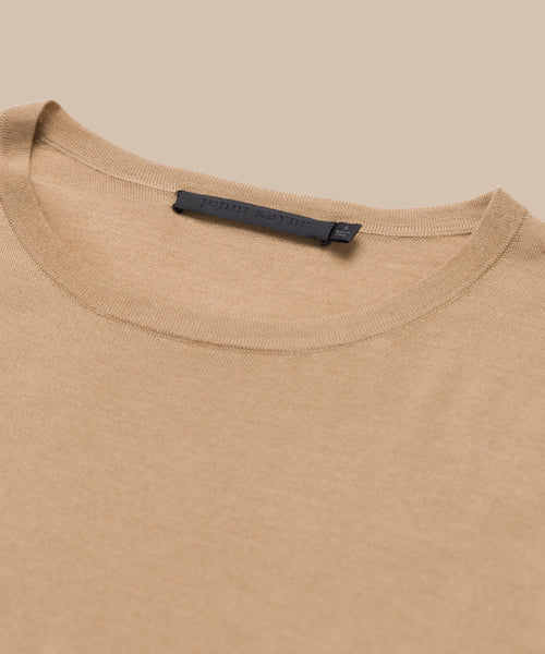 A close-up of the Mercer Crewneck shows its beige color, classic cut neckline, and a black label with text inside the collar as it lies flat.
