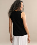A woman with long, curly hair smiles as she stands turned away, wearing the Merino Tank in black and a white skirt, set against a plain beige background.