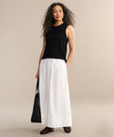 A woman with long curly hair wears the Merino Tank as a base layer, styled with a long white skirt, black bag, and black shoes, standing against a plain beige background.