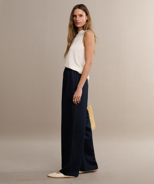 A woman stands sideways against a neutral background, wearing the Merino Tank in white, loose navy pants, cream flats, and holding a beige woven handbag.