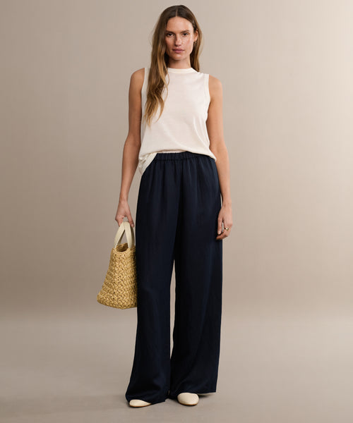 A woman stands against a beige background, wearing the Merino Tank, loose navy trousers, and white shoes. She holds a woven beige handbag and has long, wavy hair.