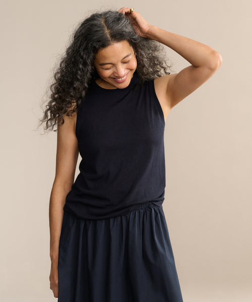 A woman with long curly hair smiles while looking down, wearing the Merino Tank in black with a matching skirt, one hand touching her hair, standing against a plain beige background.