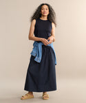A woman with long curly hair wears the Merino Tank, a long black skirt, tan sandals, and a denim jacket tied at her waist, standing against a plain beige background.