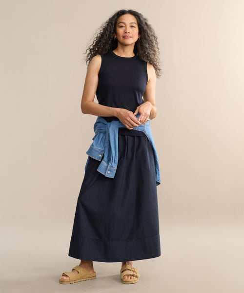 A woman with long curly hair wears the Merino Tank, a long black skirt, tan sandals, and a denim jacket tied at her waist, standing against a plain beige background.