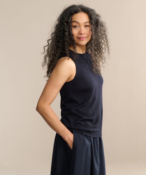 A woman with long, curly gray hair stands sideways against a neutral background, smiling softly at the camera. She wears a sleeveless black Merino Tank and a dark skirt, with one hand in her pocket.