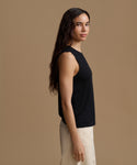 A woman in side profile wearing a black sleeveless top and light beige pants, standing against a solid warm brown studio background.