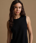 A woman wearing a black sleeveless top, photographed against a solid warm beige studio background.