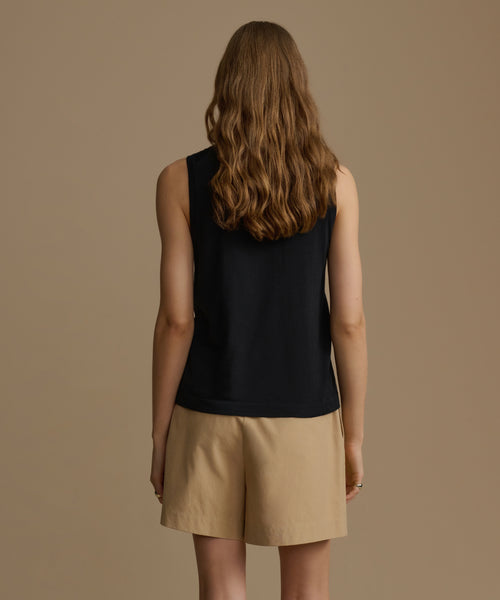 A woman shown from the back wearing a sleeveless black top and beige shorts against a plain warm taupe studio background.